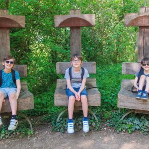 3 boys each sitting on a wooden chair in the middle of wooded area. The chairs are all beside each other in a row.